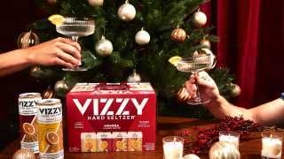 A Vizzy Mimosa Hard Seltzer case, two glasses cheers-ing in front of a Christmas tree