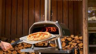 Sliding a pizza out of the Ooni Karu 12G pizza oven in front of a woodpile