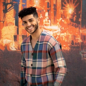 Mena Massoud smiling and standing in front of a colourful wall