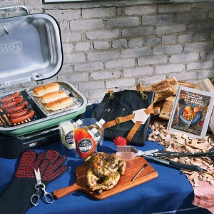 A Weber grill with hot dogs and assorted grilling accessories on a picnic table