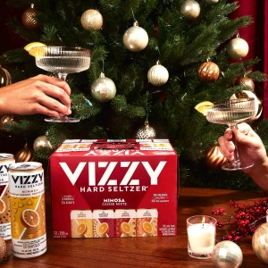 Two hands holding glasses with a case and cans of Vizzy Mimosa Hard Seltzer